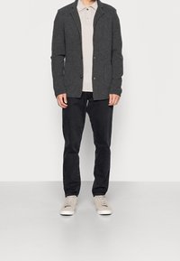 Grå texturerad cardigan med en figursydd design, två knappar och snedskuren krage, lagd över en ljusbeige polo, i kombination med svarta byxor och ljusa sneakers.