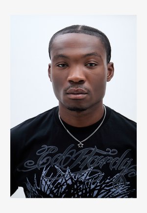 Camiseta negra con diseño gráfico en azul claro, que presenta un collar con cadena de plata y un colgante circular. Primer plano de la parte superior del cuerpo.