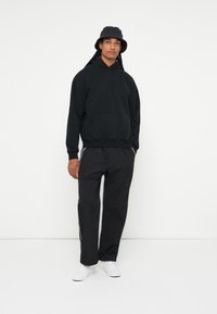 Črna kapuca s kengurujskim žepom, v kombinaciji s črnimi trenirka hlačami z belimi stranskimi črtami in belimi supergami. Krožna vedra kapa.