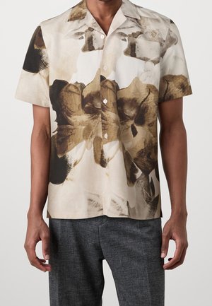 Homme portant une chemise à manches courtes boutonnée avec un grand motif floral abstrait en beige, marron et noir, associée à un pantalon gris.