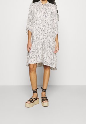 Femme portant une robe ample à motifs longueur genoux et des sandales plateformes noires avec brides à la cheville sur un fond uni.