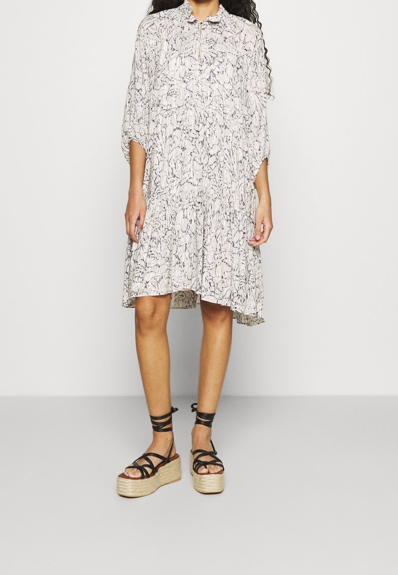 Femme portant une robe ample à motifs longueur genoux et des sandales plateformes noires avec brides à la cheville sur un fond uni.