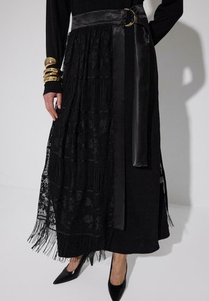 Sort maxinederdel med lagdelt blonder, frynser, et bredt satinbælte med guldsænde, kombineret med sorte spidse hæle og guldbangles om det ene håndled.