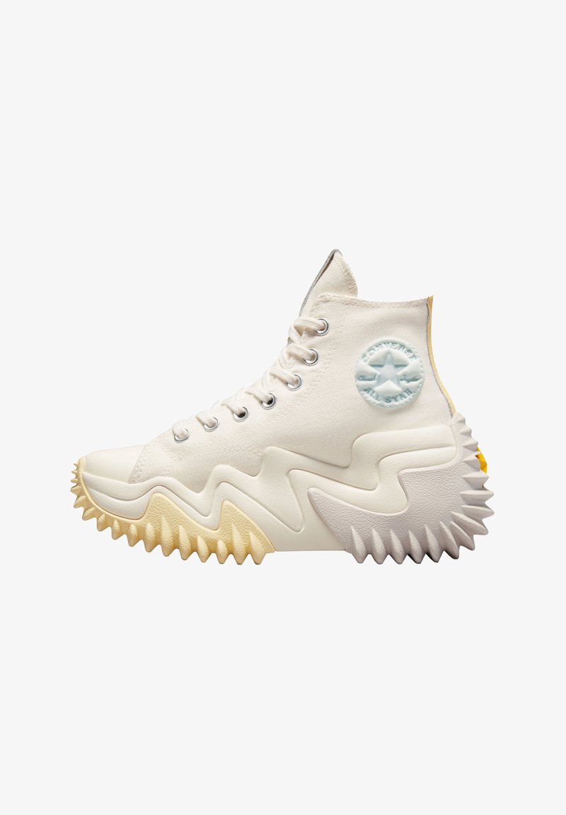 High-top sneaker in white canvas, featuring a jagged rubber sole in cream and yellow, with a blue star logo on the side and silver eyelets.