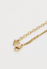 Collana in tonalità oro con un pendente a forma di triangolo, anelli a catena e chiusura a gancio. Materiali texturizzati e lucidi con una finitura brillante.