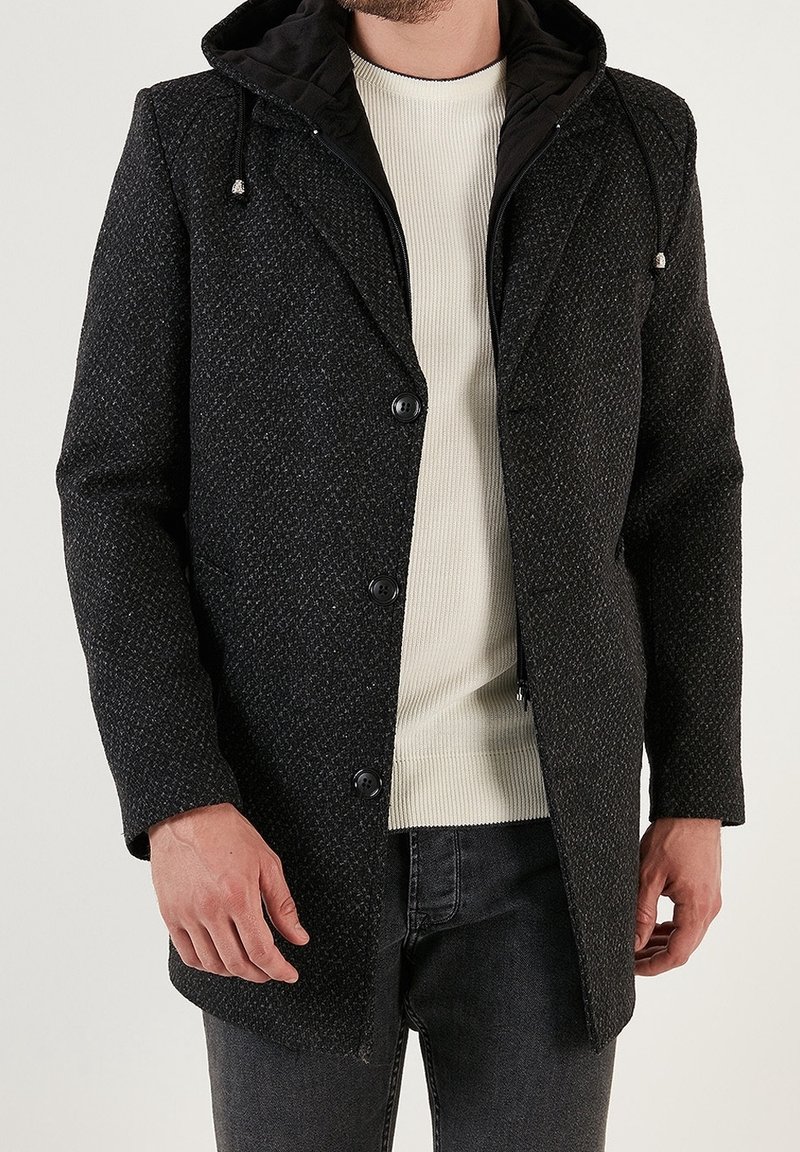 Abrigo oscuro con textura y capucha, con cierre frontal de botones y tela interior de contraste. Diseño sencillo, adecuado para uso casual.