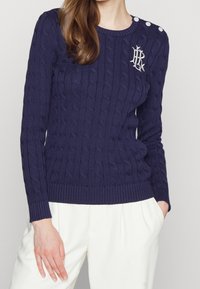 Mörkblå kabelstickad tröja med rund hals, ribbad nederkant och axelknappar. Broderad logotypdetalj på vänster bröst. Mediumviktigt tyg.