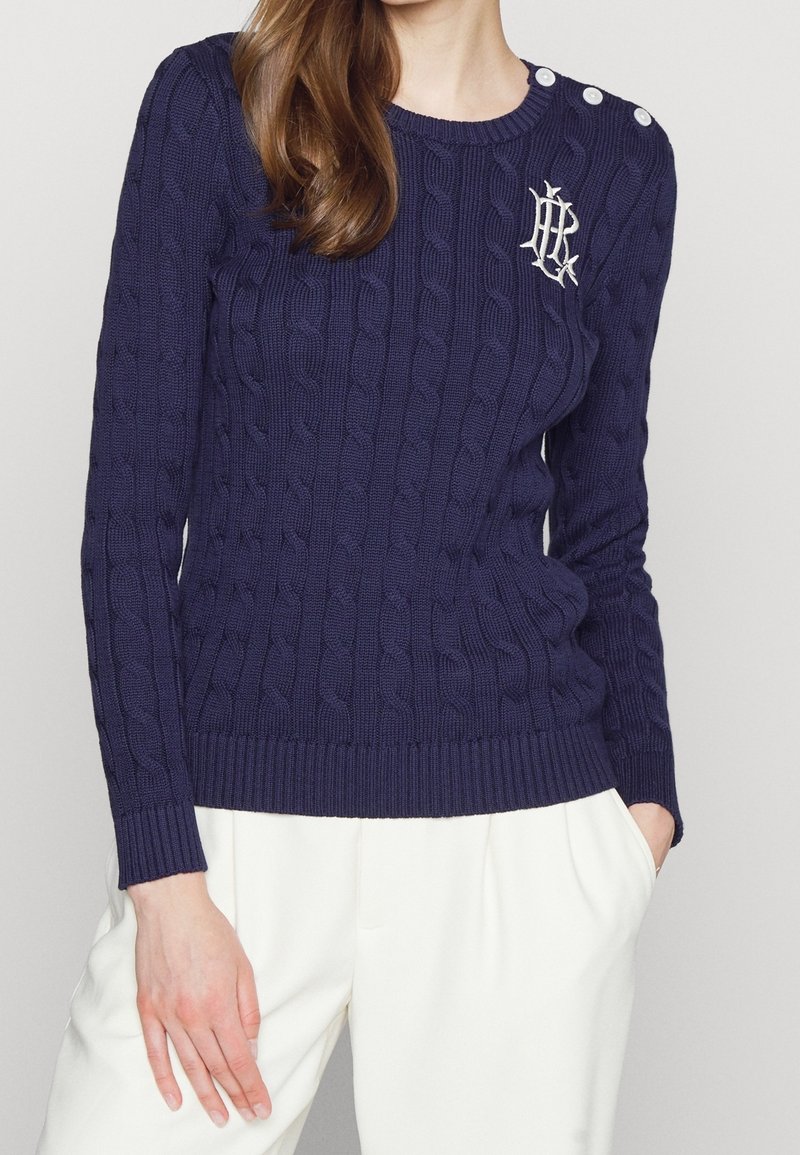 Mörkblå kabelstickad tröja med rund hals, ribbad nederkant och axelknappar. Broderad logotypdetalj på vänster bröst. Mediumviktigt tyg.