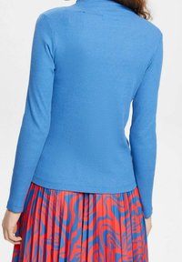 Långärmad ribbad topp i livligt blått, med hög krage och en smal passform, tillsammans med en röd mönstrad kjol.