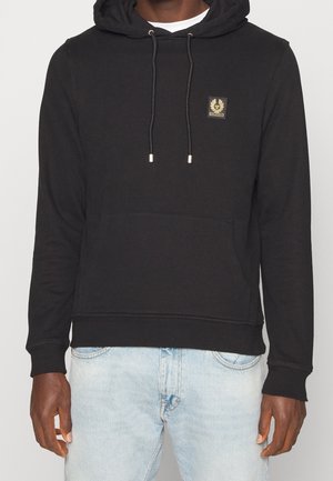 Mann trägt schwarzen Kapuzenpullover mit Vordertasche und Emblem auf der Brust, kombiniert mit hellblauen Jeans, steht vor einfachem Hintergrund.