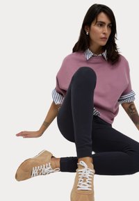 Overmaatse lila sweatshirt over een blauw-wit gestreept shirt, gecombineerd met zwarte leggings en beige sneakers met witte veters.