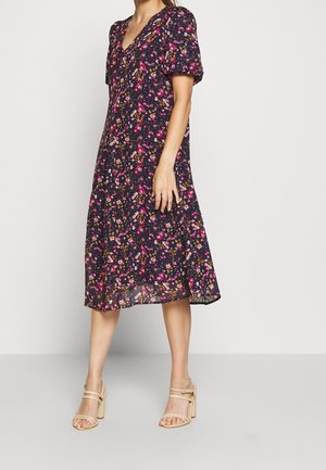 Sukienka midi w kwiatowy wzór w kolorze granatowym, z krótkimi bufiastymi rękawami, dekoltem w serek i rozkloszowanym dołem. Zestawiona z beżowymi sandałami na słupku.