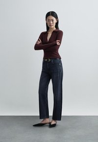 Μπορντό πλεκτό πουλόβερ με V λαιμόκοψη, συνδυασμένο με σκούρα μπλε τζιν κανονικής γραμμής και μαύρα φλατ παπούτσια, με απλή ζώνη και απαλή υφή.