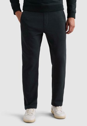 Homme portant un pantalon décontracté noir et des baskets blanches, debout avec une main dans la poche devant un fond clair uni.