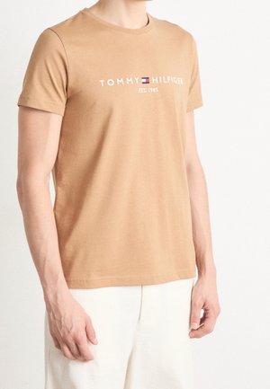 Mężczyzna w beżowej koszulce Tommy Hilfiger z krótkim rękawem z białym napisem i jasnych spodniach na białym tle.