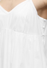 Λευκό αμάνικο φόρεμα με V λαιμόκοψη, στη μέση σούρα και κουμπωτή λεπτομέρεια στην πλάτη στο άνω μέρος του κορμού ενός ατόμου.