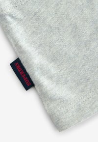 Ljust grått, mjukt tyg med en slät yta. Har en marinblå tygetikett med "SUPERDRY" i röd färg. Rak fåll med minimala sömnadsdetaljer.