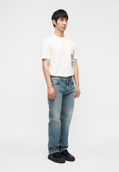 Νεαρός άνδρας στέκεται φορώντας λευκό μπλουζάκι με στάμπα, ανοιχτόχρωμο τζιν παντελόνι και μαύρα αθλητικά παπούτσια, μπροστά σε μονόχρωμο γκρι φόντο.