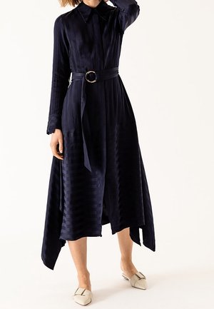 Kobieta nosząca granatową sukienkę midi z długim rękawem, paskiem w talii i asymetrycznym dołem, zestawioną z beżowymi wsuwanymi butami ze szpiczastym noskiem.