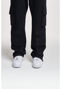 Pantalon cargo noir avec plusieurs poches, coupe ample et ourlets retroussés, associé à des baskets blanches avec des détails perforés.
