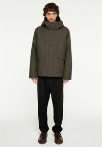 Olivgrön isolerad jacka med hög krage och huva, som har två framlitter och är knäppt med svarta knappar, bärs över svarta byxor.