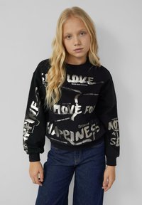 Blondynka z długimi falującymi włosami nosi czarną bluzę z białym motywacyjnym tekstem i niebieskie dżinsy, stojąc na tle jednolitego szarego tła.