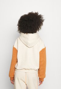 Felpa con cappuccio in tessuto beige con maniche in sherpa arancione. Orlo e polsini elasticizzati, cappuccio senza cuciture e dettagli di cucitura a contrasto.