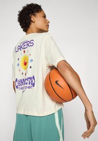 Kremo spalvos marškinėliai su "LOS ANGELES LAKERS" užrašu ir gėlių grafika. Derinami su teal spalvos šortais su baltais šoniniais dryžiais ir laikomi oranžinės spalvos krepšiniu.
