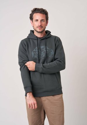 Man met kort donker haar die een donkere hoodie met een autografiek draagt en één mouw aanpast tegen een effen lichte achtergrond.