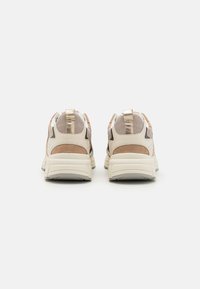 Beige sneakers med metalliska detaljer, suède och mesh-funktioner, upphöjda hälflikar och tjocka sålar. Bakifrån med fokus på distinkta texturer.