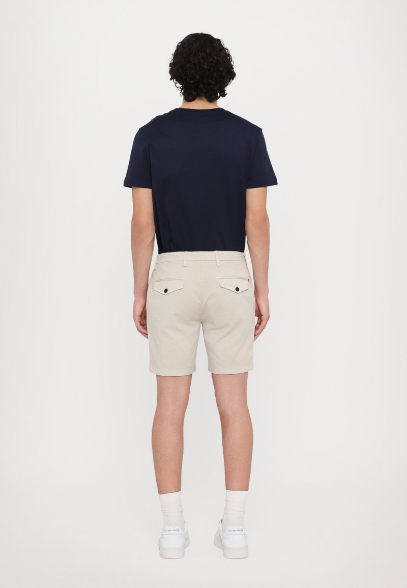 T-shirt navy, pantaloni corti beige, due tasche posteriori con bottoni neri e sneakers bianche con calzini visibili alla caviglia. Completo semplice e informale.