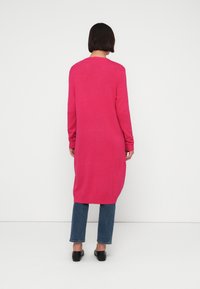 Μακριά, ροζ πλεκτή μπλούζα με μα κριά μανίκια και στρογγυλή λαιμόκοψη. Συνοδεύεται από μπλε τζιν και μαύρα παπούτσια. Απλή υφή.