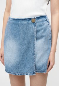 Γυναίκα που φοράει ανοιχτό μπλε μίνι φούστα από τζιν με κουμπιά μπροστά και λευκό αμάνικο τοπ, στέκεται μπροστά σε απλό φόντο.