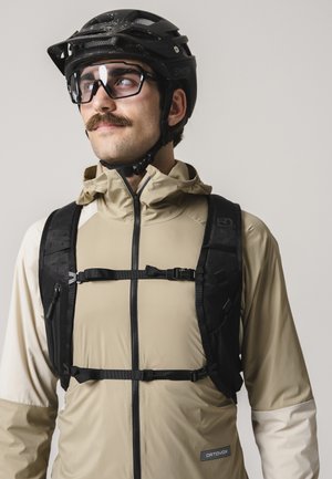 Mann trägt beige Jacke, schwarzen Fahrradhelm, Schutzbrille und schwarzen Brust-Rucksack und schaut leicht seitlich vor einfachem Hintergrund.