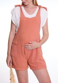 Pfirsichfarbene Leinenlatzhosen mit verstellbaren Bindeträgen, Seitentaschen und umgeschlagenem Saum, getragen über einem weißen V-Ausschnitt-Shirt.