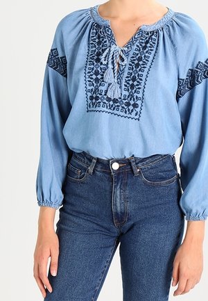 Persona con una blusa azul bordada con lazos y flecos y unos vaqueros azul oscuro de talle alto, de pie con los brazos relajados a los lados.