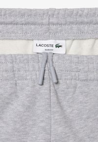 Pantaloni de trening subțiri, de culoare gri deschis, cu talie elastică, șnururi și eticheta cu logo-ul Lacoste. Textură moale și design simplu.