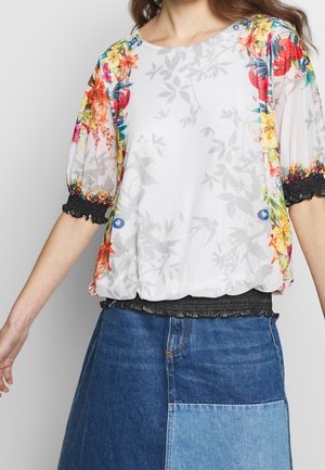 Kvinde iført hvid bluse med farverigt blomsterprint på puffede ærmer og rynkede sorte elastiske manchetter, kombineret med tofarvet denimnederdel.