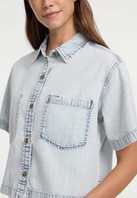 Helles blaues Denim-Hemd mit kurzen Ärmeln, Knopfleiste, Brusttasche und kontrastierenden Nähten an Kragen und Taschenkanten.