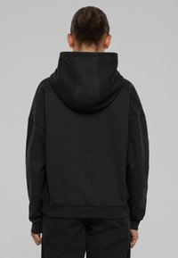 Persona con el cabello recogido en un moño, vistiendo un hoodie negro holgado y pantalones a juego, de pie frente a un fondo liso gris claro.