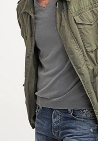 Oliwkowa kurtka w stylu militarnym z wieloma kieszeniami, nałożona na szary, dopasowany sweter z dekoltem w serek oraz niebieskie, postrzępione dżinsy.