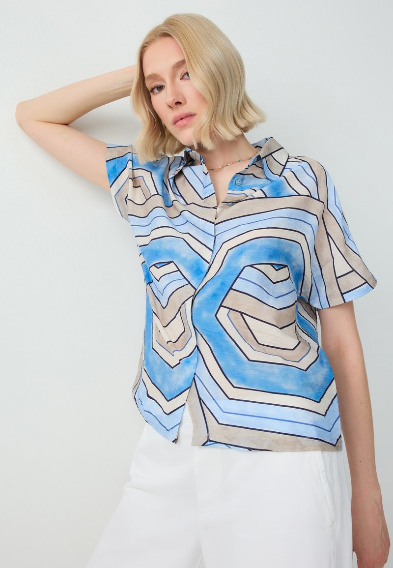 Femme blonde portant une chemise boutonnée à manches courtes à motifs bleu, beige et noir avec un pantalon blanc, posant avec un bras levé.