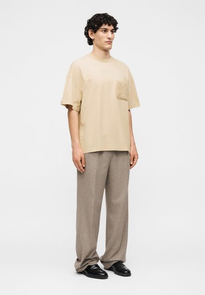 Νέος άνδρας με σγουρά μαλλιά που φοράει μπεζ υπερμεγέθη μπλούζα με υφή τσέπης, χαλαρό παντελόνι ταρπ και μαύρα δερμάτινα παπούτσια, στέκεται ακίνητος.