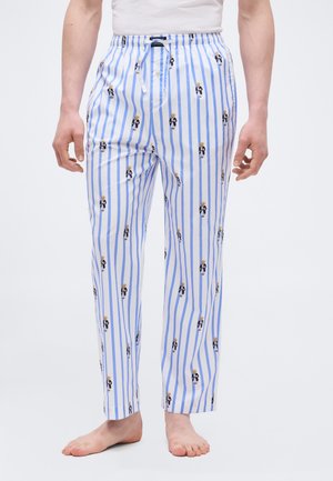 Weiße Herren-Pyjamahose mit vertikalen blauen Streifen und wiederholten Prints eines Bären im Anzug, barfuß getragen mit einem weißen, in die Hose gesteckten Hemd.