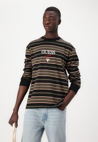 Camisa negra de manga larga con rayas horizontales en verde, rosa y beige; presenta el logotipo "GUESS" bordado en blanco y rojo en el pecho.