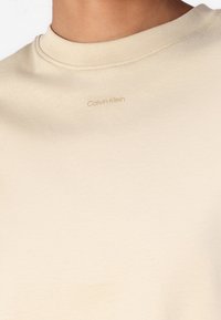 Beige cotton t-shirt with a crew neckline and short sleeves. Features a subtle “Calvin Klein” logo in matching color on the chest.