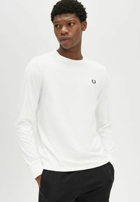 T-shirt bianco in cotone a maniche lunghe con scollatura rotonda e un piccolo emblema nero sul lato sinistro del petto. Tessuto morbido, vestibilità casual.
