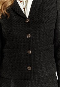 Blazer nero con texture, colletto a scialle, bottoni floreali dorati e due tasche frontali. Il tessuto presenta un motivo in rilievo.