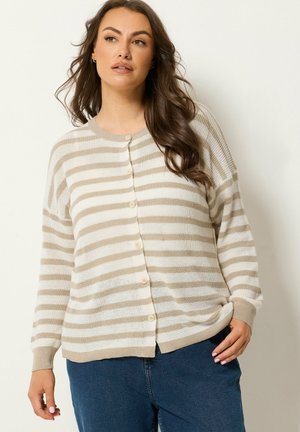Kvinde iført beige og hvide stribede knap-op cardigan med lange ærmer og blå jeans mod en ensfarvet baggrund.