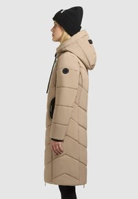 Beige pufferjacka med huva, med chevron-quilting, sidofickor och svarta detaljer. Bärs med en svart stickad mössa.